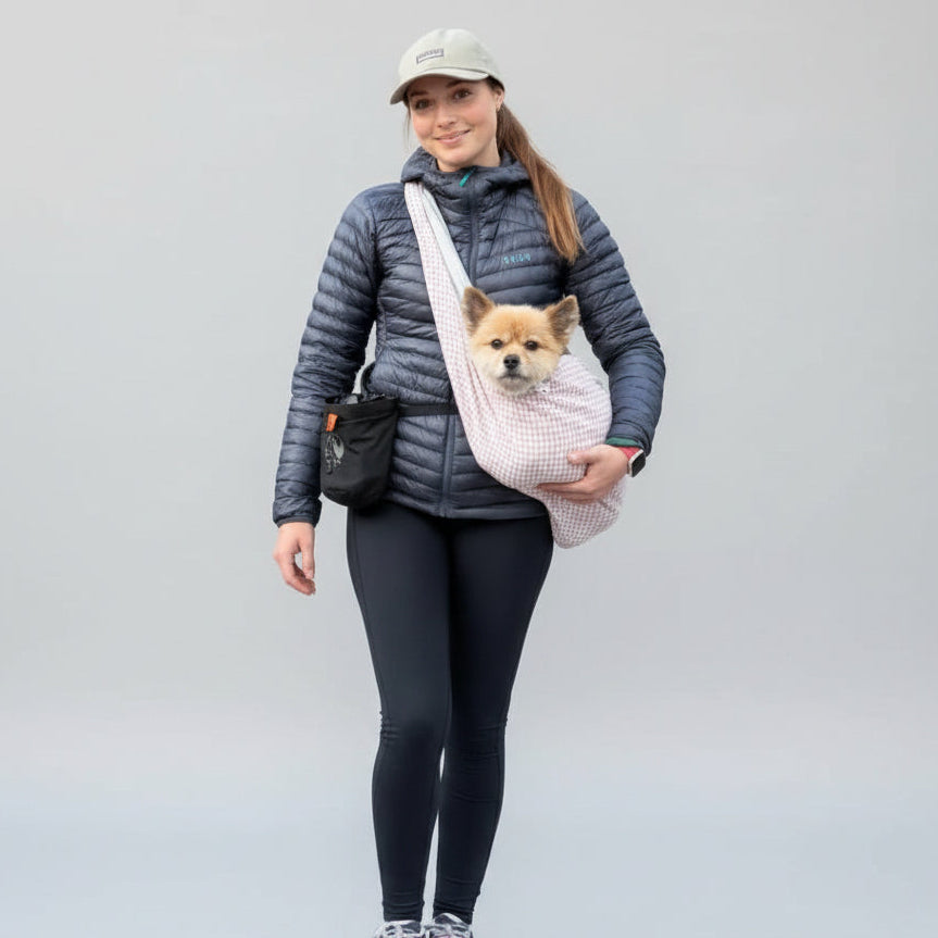 A woman walking on a tree-lined road wearing a light pink pet carrier bag across her chest with a small dog inside. She is dressed in a dark puffer jacket, black leggings, and pink sneakers.