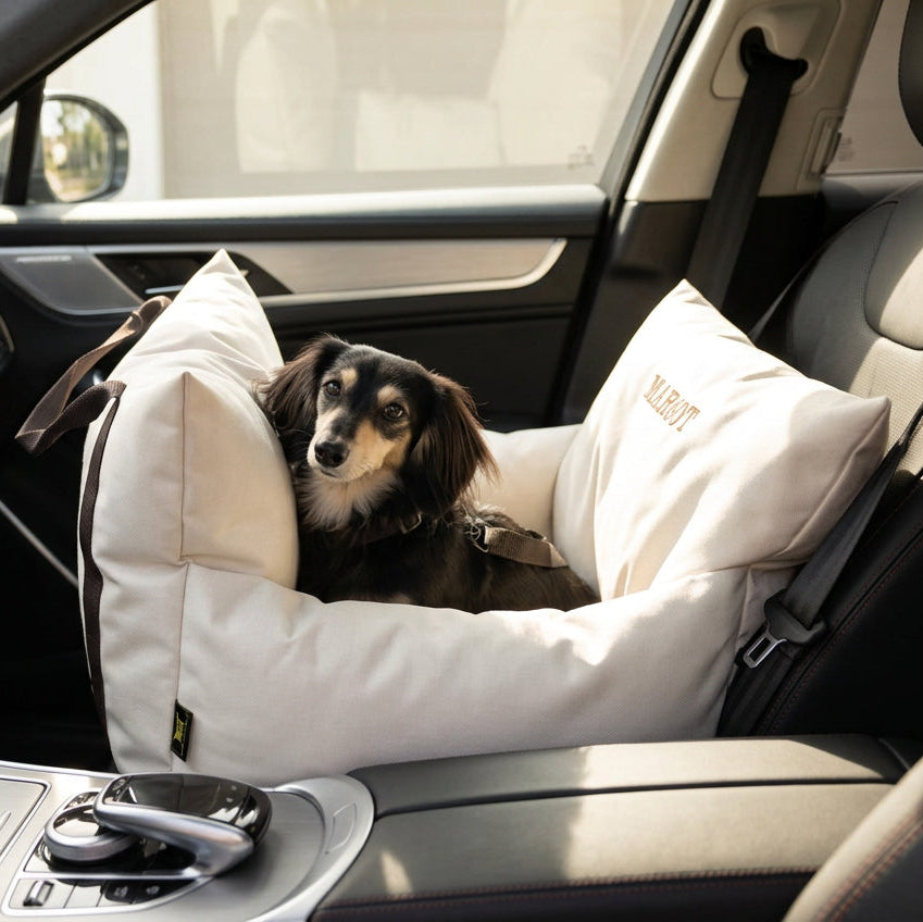 A luxury dog car seat in stone color with a black dog peeking out from the cushion. The seat is installed in a vehicle and has a waterproof exterior.