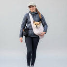 A woman walking on a tree-lined road wearing a light pink pet carrier bag across her chest with a small dog inside. She is dressed in a dark puffer jacket, black leggings, and pink sneakers.