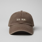 A brown washed cotton baseball cap with 'DOG MUM' embroidered in beige on the front, resting on a white desk with papers and cables visible in the background.