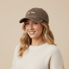 A brown washed cotton baseball cap with 'DOG MUM' embroidered in beige on the front, resting on a white desk with papers and cables visible in the background.
